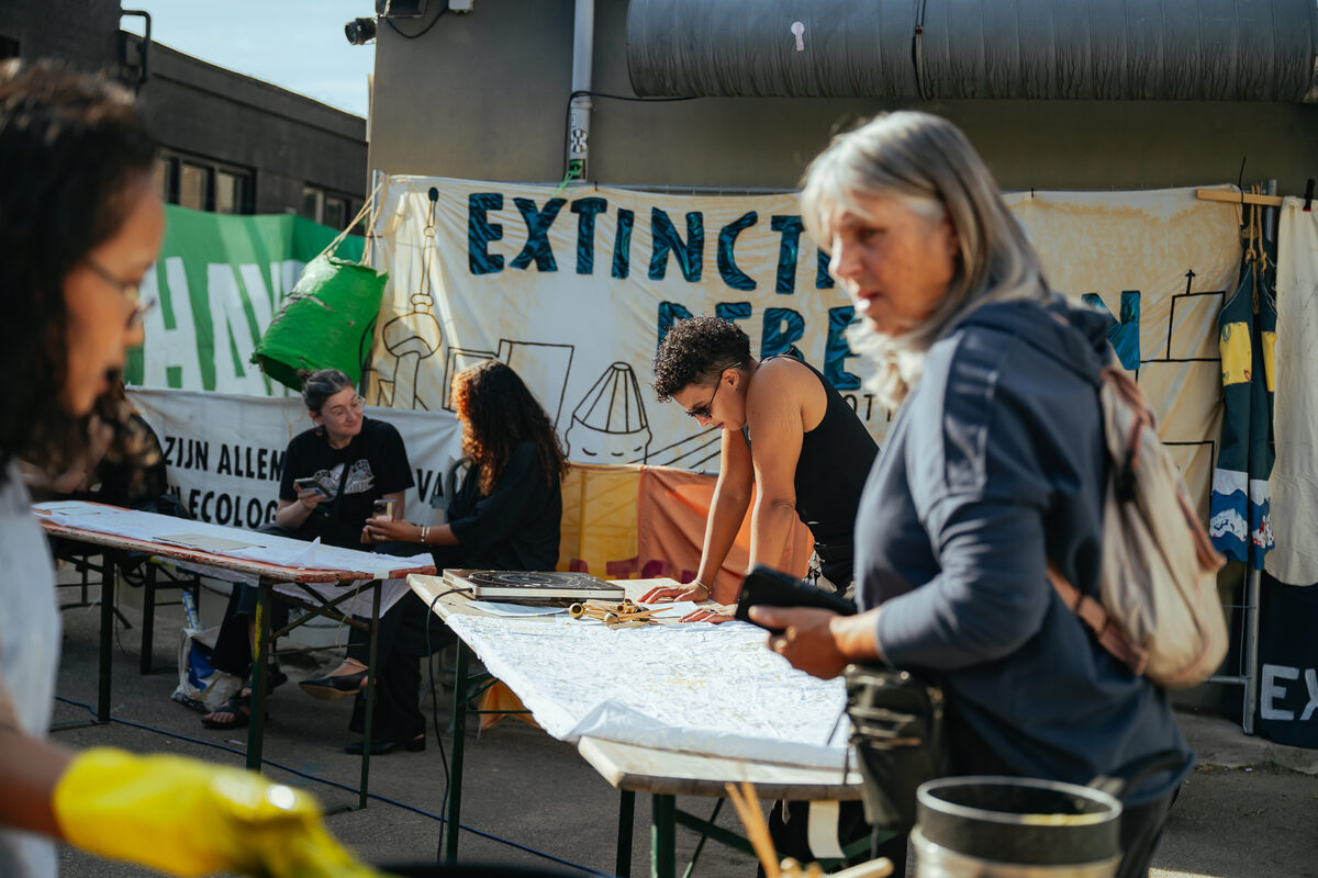 Participants drawing batik at long outdoor tables in the Brutus yard, protest banners strung behind them