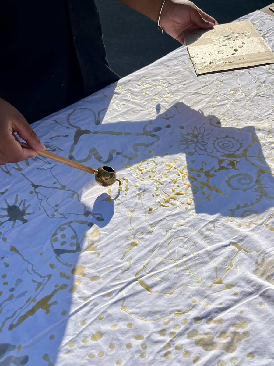 A hand drawing hot wax onto white cloth with a brass tjanting