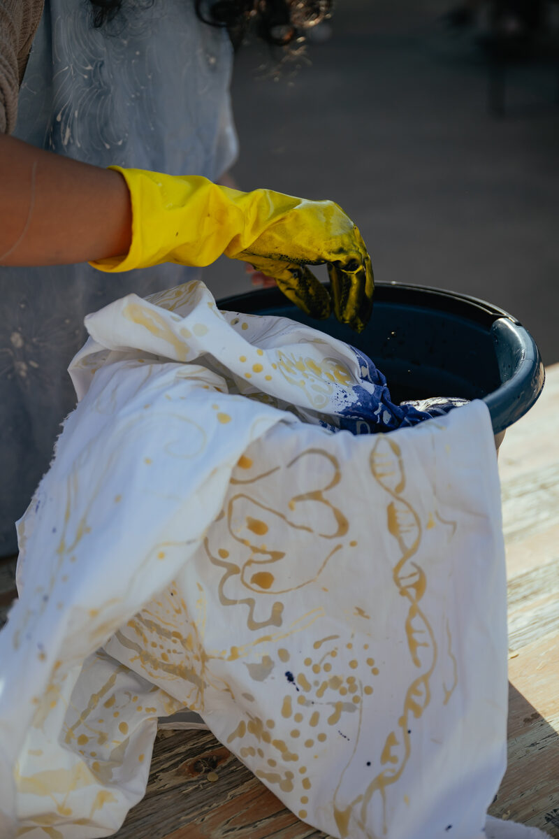 A gloved hand lowering the waxed cloth into a bucket of indigo dye